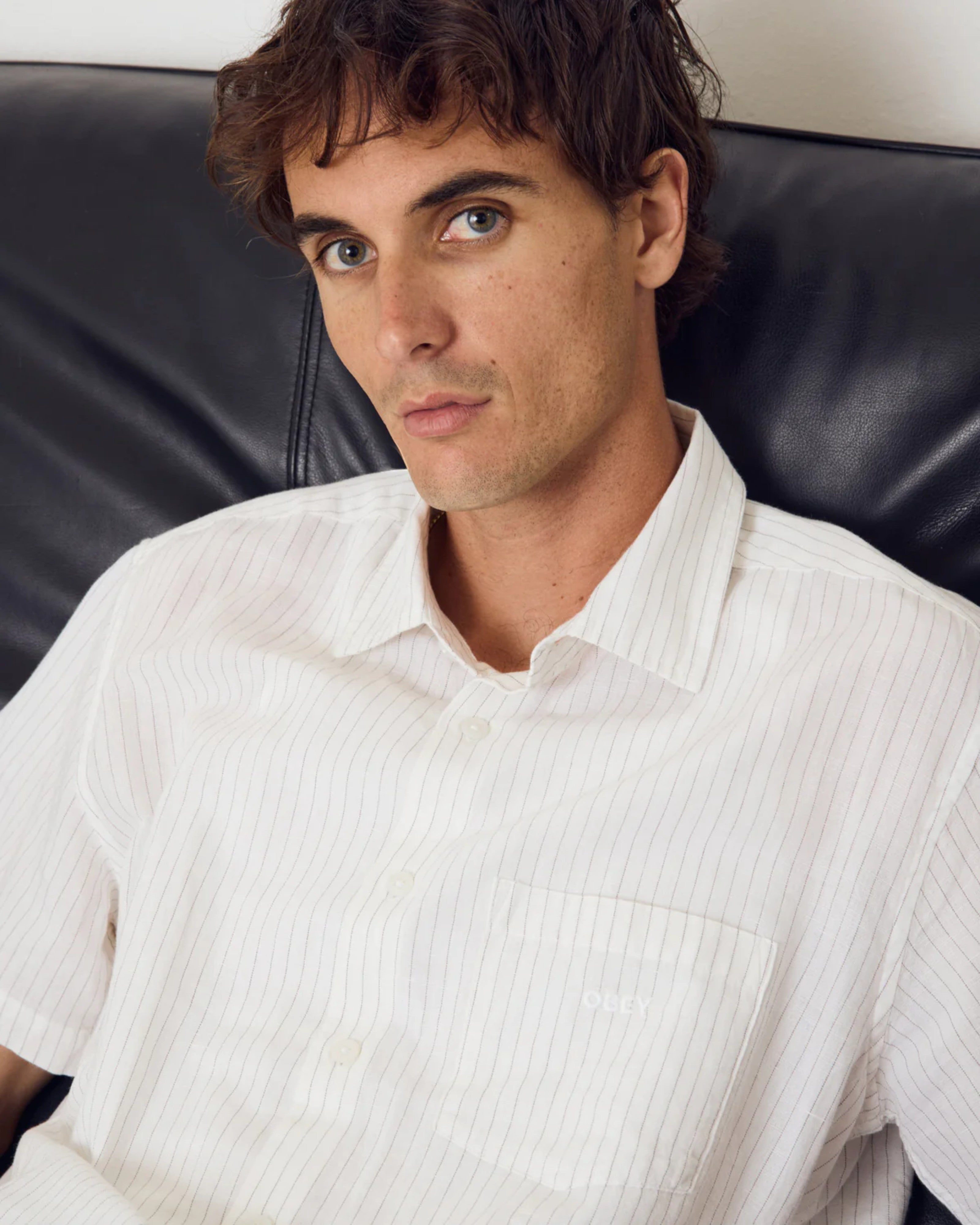 Astrofat Obey Barry Woven short-sleeve white button-up shirt with pinstripes and a chest pocket. Model is seated on a black leather couch.