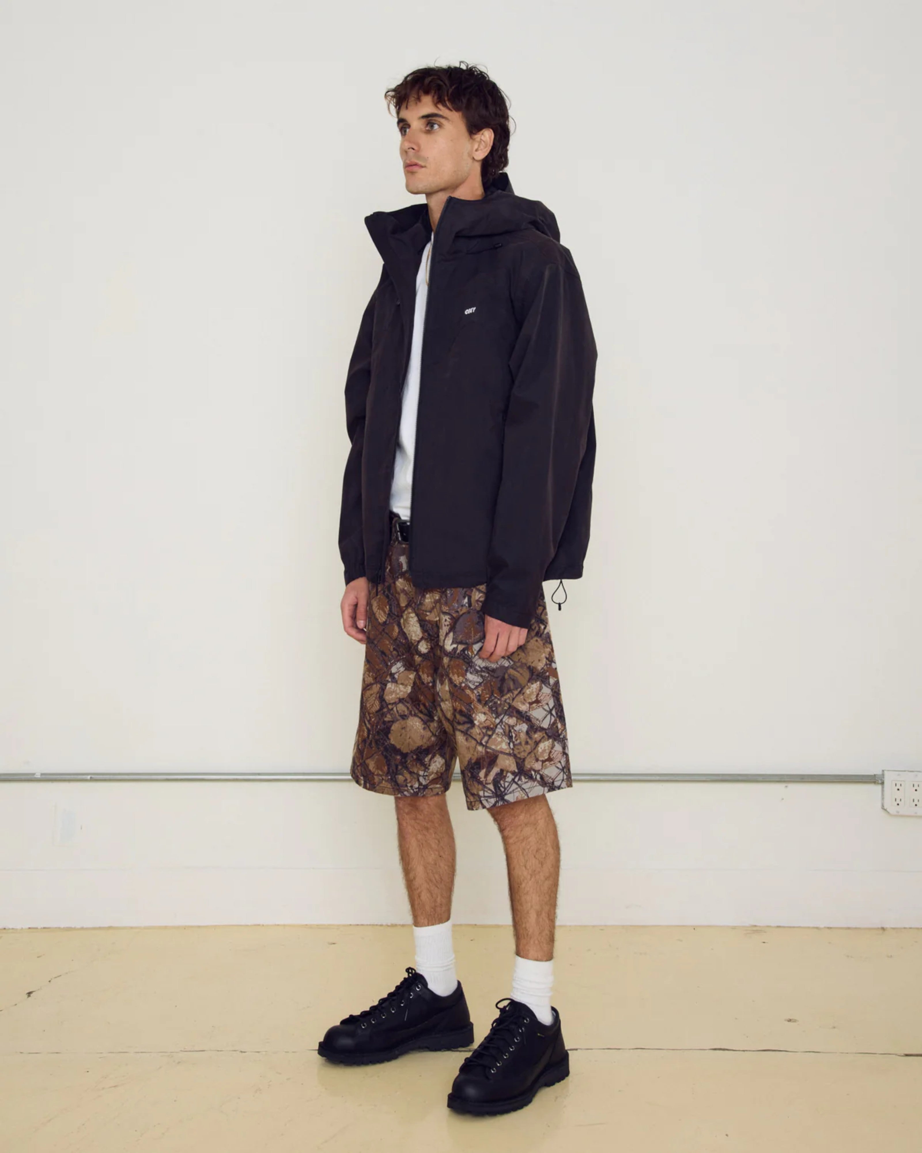Astrofat Obey Sota Shell Jacket in black, featuring a relaxed fit, adjustable hood, zip closure, and embroidered logo. Model wears it with camouflage shorts and black shoes.