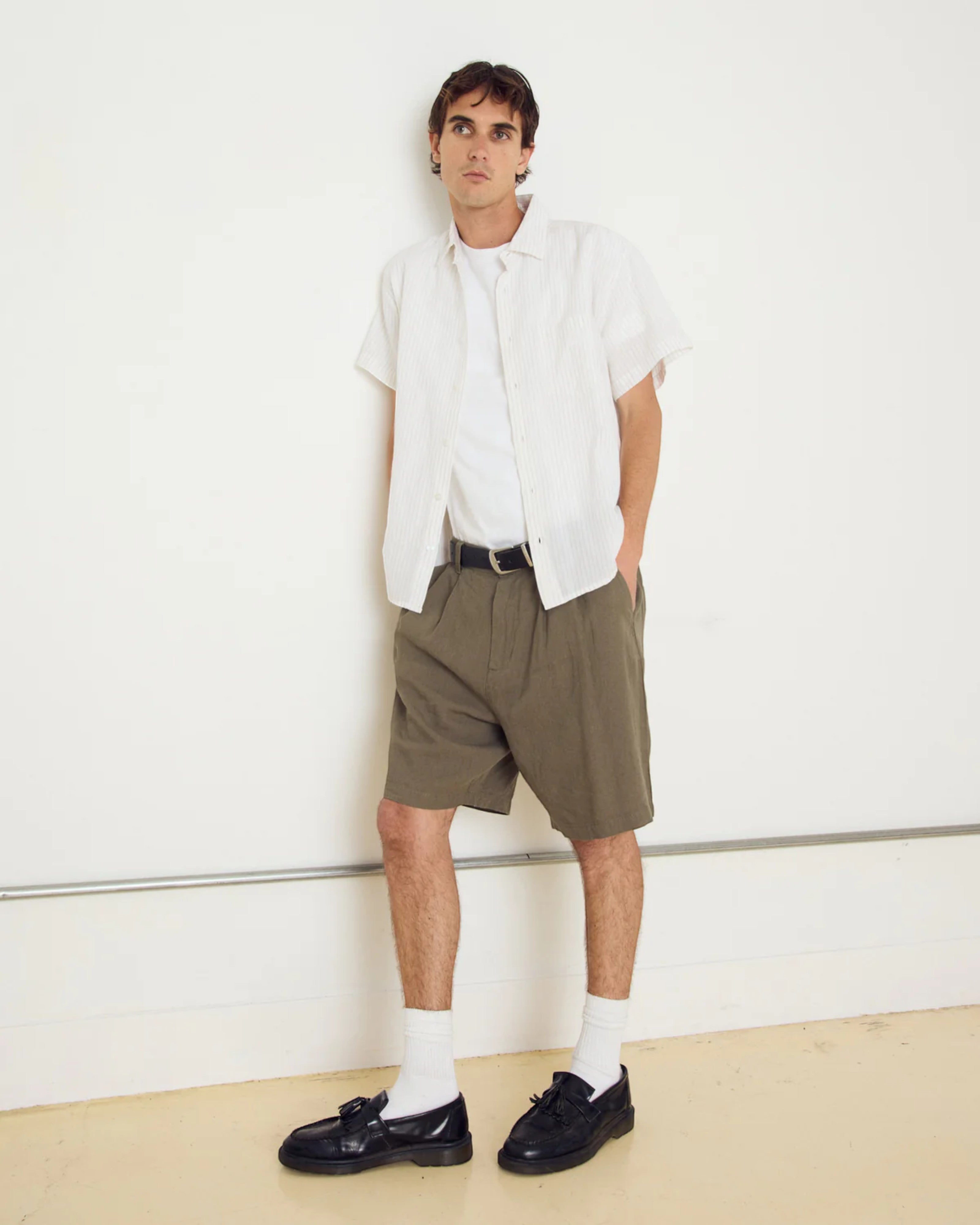 Astrofat model wears a white short-sleeve button-up shirt with subtle vertical stripes over a white t-shirt, paired with olive green shorts and black loafers.